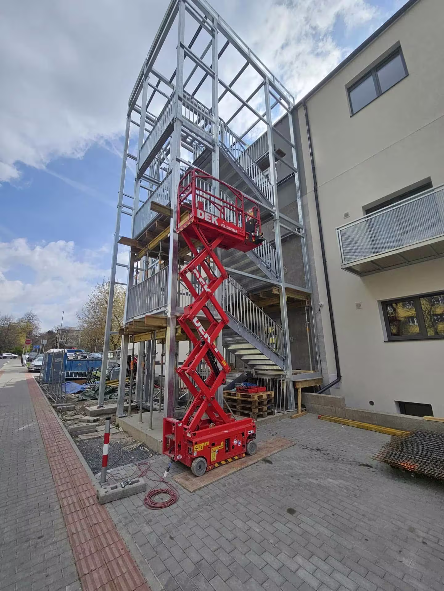 Použití červené nůžkové plošiny při montáži ocelové konstrukce externí výtahové šachty u panelového domu.